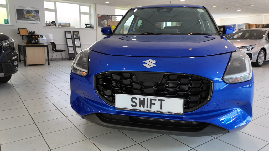 The front of the Suzuki Swift 2024 with a redesigned grille, L-shaped LED daytime running lights, and a badge on the hood.